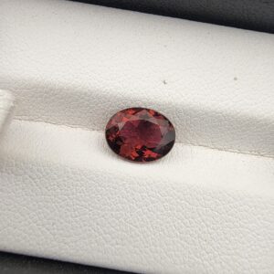 ​A rare 1.69-carat natural Mozambique Tourmaline gemstone. In the first image, it is held securely in silver metal tweezers against a clean gray background, showcasing a vibrant plum-purple core with bright orangish-amber flashes around its faceted edges. The second image shows the loose oval gemstone resting flat on a white textured leather insert inside a display box, emphasizing its deep, velvety wine-red and purple body tones.