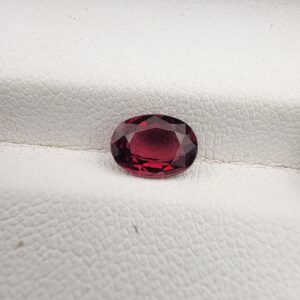 ​A premium 1.15-carat natural Mozambique Rubellite Tourmaline gemstone resting flat on a white textured leather insert inside a display box. The loose oval faceted gemstone showcases an exceptionally rich, deep crimson and magenta-red body color with crisp, light-reflecting crown facets.