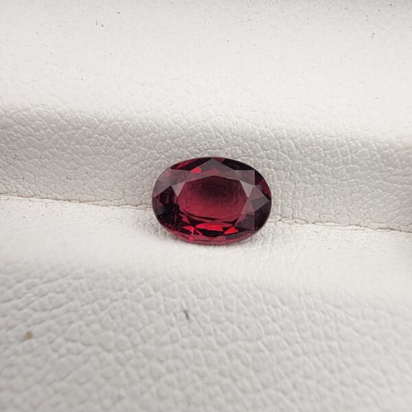 ​A premium 1.15-carat natural Mozambique Rubellite Tourmaline gemstone resting flat on a white textured leather insert inside a display box. The loose oval faceted gemstone showcases an exceptionally rich, deep crimson and magenta-red body color with crisp, light-reflecting crown facets.