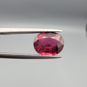 A premium 1.67-carat natural Mozambique Rubellite Tourmaline gemstone. In the first image, it is held securely in silver metal tweezers against a clean gray background, flashing a vibrant magenta-red and showcasing beautiful natural internal inclusion patterns under direct light. The second image shows the loose oval faceted gemstone resting flat on a white textured leather insert inside a display box, highlighting its rich, deep crimson body color.