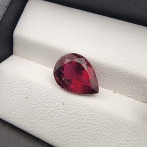 ​A premium 3.41-carat natural Mozambique Rubellite Tourmaline gemstone resting elegantly on a white textured leather insert inside a black presentation box. The loose, earth-mined gemstone is precision faceted into a beautifully proportioned pear shape, exhibiting a deeply saturated magenta-red body color with rich wine-red flashes and showcasing intricate, beautiful internal inclusions reflecting from its teardrop crown.