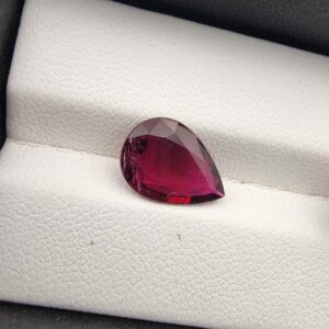 A premium 2.64-carat natural Mozambique Rubellite Tourmaline gemstone. In the first image, the stone is photographed in a close-up shot resting on a white textured leather display insert, capturing its vibrant, glowing magenta-red heart and delicate natural internal inclusions. The second image provides a slightly wider angle of the faceted pear-shaped gemstone inside its black display box, highlighting its rich, deep wine-red body color and symmetrical teardrop silhouette.
