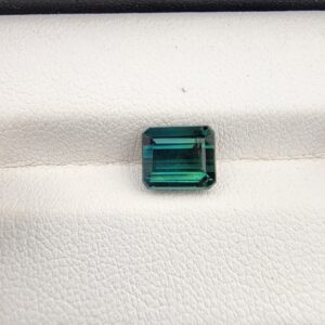 ​A premium 2.35-carat natural African Greenish-Blue Tourmaline gemstone shown across two views. In the first image, the stone is held securely in silver metal tweezers against a clean gray background, flashing intense teal-blue and minty sea-green light from its crisp pavilion facets. The second image shows the loose octagon-cut gemstone resting flat on a white textured display insert inside a black box, highlighting its rich, deep blue-green body color.