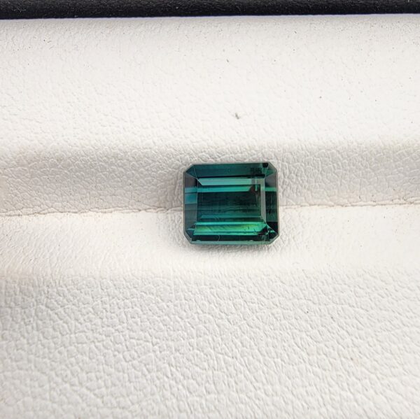 ​A premium 2.35-carat natural African Greenish-Blue Tourmaline gemstone shown across two views. In the first image, the stone is held securely in silver metal tweezers against a clean gray background, flashing intense teal-blue and minty sea-green light from its crisp pavilion facets. The second image shows the loose octagon-cut gemstone resting flat on a white textured display insert inside a black box, highlighting its rich, deep blue-green body color.