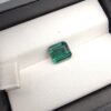 ​A premium 2.29-carat natural African Greenish-Blue Tourmaline gemstone shown across two views. In the first image, the stone is held securely in silver metal tweezers against a clean gray background, flashing intense teal-blue and minty sea-green light from its crisp pavilion facets. The second image shows the loose octagon-cut gemstone resting flat on a white textured display insert inside a black box, highlighting its rich, deep blue-green body color.