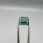 ​A premium 2.29-carat natural African Greenish-Blue Tourmaline gemstone shown across two views. In the first image, the stone is held securely in silver metal tweezers against a clean gray background, flashing intense teal-blue and minty sea-green light from its crisp pavilion facets. The second image shows the loose octagon-cut gemstone resting flat on a white textured display insert inside a black box, highlighting its rich, deep blue-green body color.