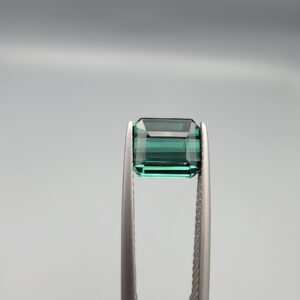 ​A premium 2.29-carat natural African Greenish-Blue Tourmaline gemstone shown across two views. In the first image, the stone is held securely in silver metal tweezers against a clean gray background, flashing intense teal-blue and minty sea-green light from its crisp pavilion facets. The second image shows the loose octagon-cut gemstone resting flat on a white textured display insert inside a black box, highlighting its rich, deep blue-green body color.