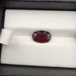 ​A premium 3.60-carat natural Madagascar Red Garnet gemstone shown across three product views. The stone is photographed held securely in silver metal tweezers against a clean gray background and resting on a white textured display insert inside a black box, showcasing its rich crimson-red body color and vibrant, fire-red sparkling facets.
