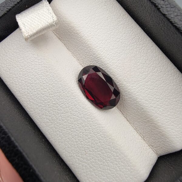 ​A premium 3.60-carat natural Madagascar Red Garnet gemstone shown across three product views. The stone is photographed held securely in silver metal tweezers against a clean gray background and resting on a white textured display insert inside a black box, showcasing its rich crimson-red body color and vibrant, fire-red sparkling facets.