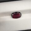 ​A premium 3.60-carat natural Madagascar Red Garnet gemstone shown across three product views. The stone is photographed held securely in silver metal tweezers against a clean gray background and resting on a white textured display insert inside a black box, showcasing its rich crimson-red body color and vibrant, fire-red sparkling facets.