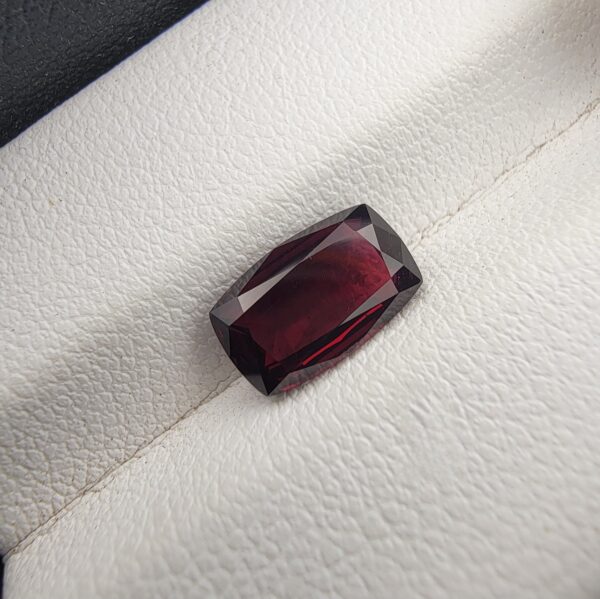 ​A premium 2.61-carat natural Madagascar Red Garnet gemstone. In the first image, it is held securely in silver metal tweezers against a clean gray background, flashing bright, intense reddish-sparkling secondary facets under direct light. The second image shows the loose cushion faceted gemstone resting flat on a white textured leather insert inside a display box, highlighting its rich, deep crimson body color.