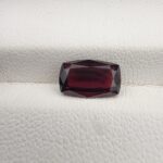 ​A premium 2.61-carat natural Madagascar Red Garnet gemstone. In the first image, it is held securely in silver metal tweezers against a clean gray background, flashing bright, intense reddish-sparkling secondary facets under direct light. The second image shows the loose cushion faceted gemstone resting flat on a white textured leather insert inside a display box, highlighting its rich, deep crimson body color.