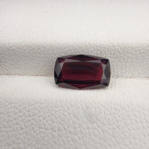 A premium 2.61-carat natural Madagascar Red Garnet gemstone. In the first image, it is held securely in silver metal tweezers against a clean gray background, flashing bright, intense reddish-sparkling secondary facets under direct light. The second image shows the loose cushion faceted gemstone resting flat on a white textured leather insert inside a display box, highlighting its rich, deep crimson body color.
