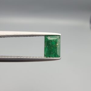 ​A rare 0.81-carat natural Swat Valley Emerald gemstone resting flat on a white textured leather insert inside a black presentation box. The loose rectangular-cut beryl showcases a vibrant, saturated deep green body color featuring highly distinct, shimmering golden pyrite inclusions locked inside its crystalline core.