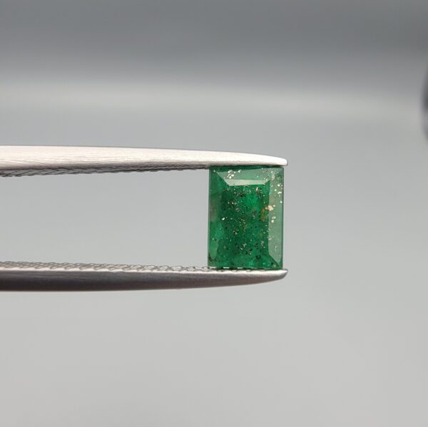 ​A rare 0.81-carat natural Swat Valley Emerald gemstone resting flat on a white textured leather insert inside a black presentation box. The loose rectangular-cut beryl showcases a vibrant, saturated deep green body color featuring highly distinct, shimmering golden pyrite inclusions locked inside its crystalline core.