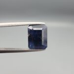 A rare 5.07-carat natural Afghan Blue Sapphire gemstone held securely in metal gemstone tweezers against a smooth gray studio background. The loose, unheated corundum features a deep, velvety midnight blue color cut into a sophisticated rectangular shape, showcasing an organic, eye-visible natural crystal growth cavity on its left-hand side.