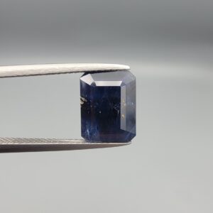 A rare 5.07-carat natural Afghan Blue Sapphire gemstone held securely in metal gemstone tweezers against a smooth gray studio background. The loose, unheated corundum features a deep, velvety midnight blue color cut into a sophisticated rectangular shape, showcasing an organic, eye-visible natural crystal growth cavity on its left-hand side.