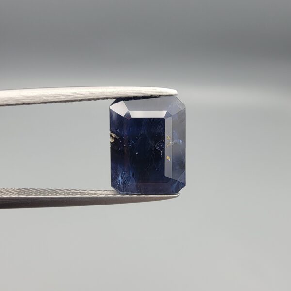 A rare 5.07-carat natural Afghan Blue Sapphire gemstone held securely in metal gemstone tweezers against a smooth gray studio background. The loose, unheated corundum features a deep, velvety midnight blue color cut into a sophisticated rectangular shape, showcasing an organic, eye-visible natural crystal growth cavity on its left-hand side.