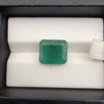 ​A premium, heavyweight 7.41-carat natural Zambian Emerald gemstone resting flat on a white textured presentation insert inside a display box. The loose, earth-mined beryl gem showcases a deeply saturated, vibrant grass-green body color with an excellent table spread and fine symmetry.