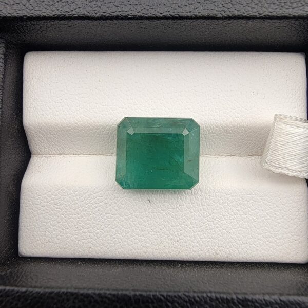 ​A premium, heavyweight 7.41-carat natural Zambian Emerald gemstone resting flat on a white textured presentation insert inside a display box. The loose, earth-mined beryl gem showcases a deeply saturated, vibrant grass-green body color with an excellent table spread and fine symmetry.