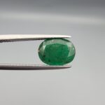 ​A premium 2.60-carat natural Swat Valley Emerald gemstone resting flat on a white textured presentation insert inside a black display box. The loose, earth-mined beryl gem is precision cut into an elegant oval shape, showcasing a vibrant grass-green body color filled with distinct, shimmering golden pyrite crystals locked beautifully inside its crystalline core.