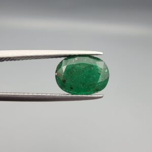 ​A premium 2.60-carat natural Swat Valley Emerald gemstone resting flat on a white textured presentation insert inside a black display box. The loose, earth-mined beryl gem is precision cut into an elegant oval shape, showcasing a vibrant grass-green body color filled with distinct, shimmering golden pyrite crystals locked beautifully inside its crystalline core.