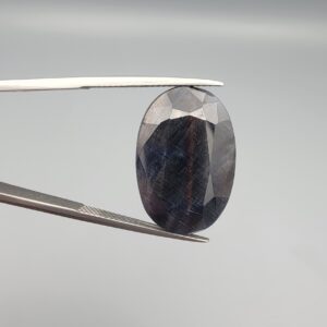 An extraordinary, investment-grade 27.59-carat natural Afghan Blue Sapphire gemstone held vertically in professional metal gemstone tweezers against a smooth, neutral gray background. The massive loose corundum gem is precision tailored into a clean octagon step-cut shape, displaying a deep, velvety midnight-blue body color accented by fine, reflective golden mineral rutile patterns and natural growth fingerprints near its surface.