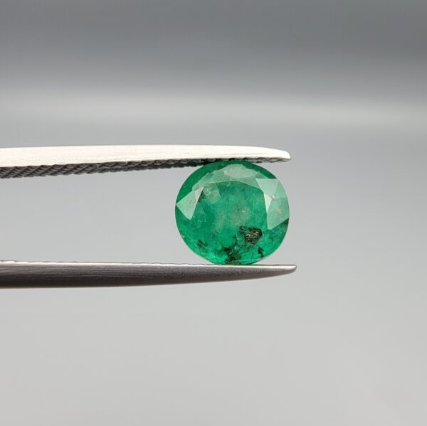 ​A rare 1.48-carat natural Swat Valley Emerald gemstone held horizontally in metal gemstone tweezers against a smooth gray studio background. The loose beryl gem features a crisp, symmetrical circular shape, exhibiting a vivid alpine-green body color accented by distinct, metallic golden pyrite crystal inclusions resting near its surface facets.