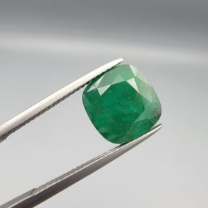 A rare and substantial 5.53-carat natural Swat Valley Emerald gemstone held securely in metal gemstone tweezers against a smooth gray background. The premium loose beryl gemstone is cut into a soft, elegant cushion shape, displaying an intense, saturated alpine-green body color with beautifully layered internal growth features.