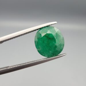 A premium 4.66-carat natural Swat Valley Emerald gemstone held securely in metal gemstone tweezers against a smooth gray studio background. The loose, earth-mined beryl gem is precision cut into an elegant, highly symmetrical circular shape, showcasing a vibrant, richly saturated green body color with intricate natural internal growth features and fine facet symmetry.
