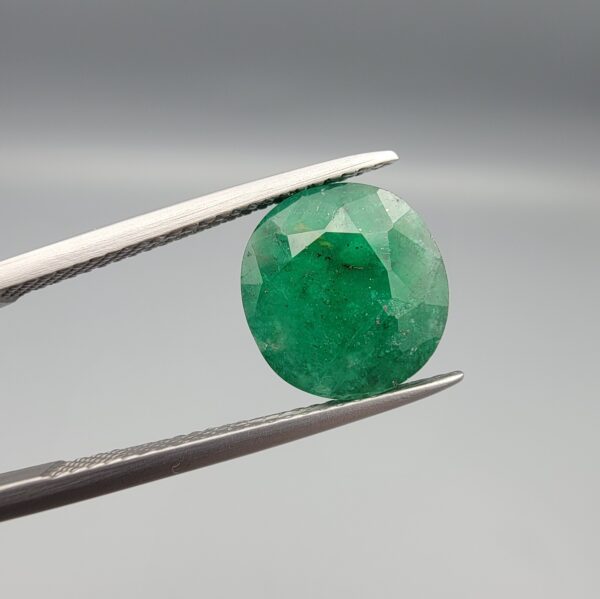 ​A premium 4.66-carat natural Swat Valley Emerald gemstone held securely in metal gemstone tweezers against a smooth gray studio background. The loose, earth-mined beryl gem is precision cut into an elegant, highly symmetrical circular shape, showcasing a vibrant, richly saturated green body color with intricate natural internal growth features and fine facet symmetry.