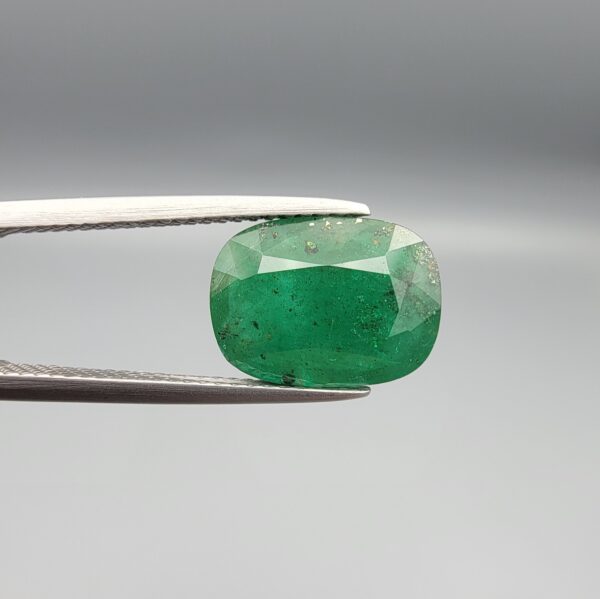​A premium 3.80-carat natural Swat Valley Emerald gemstone held securely in metal gemstone tweezers against a smooth gray studio background. The loose, earth-mined beryl gem is precision cut into an elegant cushion shape, displaying an intense, saturated alpine-green body color with beautifully layered internal growth features and fine facet symmetry.