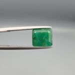 ​A premium 3.67-carat natural Swat Valley Emerald gemstone held securely in metal gemstone tweezers against a smooth gray studio background. The loose, earth-mined beryl gem is precision cut into an elegant octagon step-cut shape, showcasing an intense, saturated alpine-green body color with beautifully layered internal growth features and pristine facet symmetry.