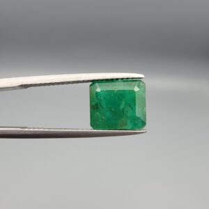 A premium 3.67-carat natural Swat Valley Emerald gemstone held securely in metal gemstone tweezers against a smooth gray studio background. The loose, earth-mined beryl gem is precision cut into an elegant octagon step-cut shape, showcasing an intense, saturated alpine-green body color with beautifully layered internal growth features and pristine facet symmetry.