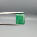 A premium 3.93-carat natural Swat Valley Emerald gemstone held horizontally in metal gemstone tweezers against a smooth gray studio background. The loose, earth-mined beryl gem is precision cut into an elegant octagon step-cut shape, showcasing an intense, saturated grass-green color with fine parallel step facets and classic internal growth features.