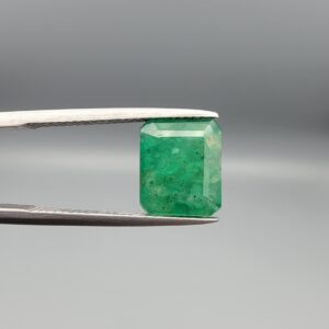 A premium 3.93-carat natural Swat Valley Emerald gemstone held horizontally in metal gemstone tweezers against a smooth gray studio background. The loose, earth-mined beryl gem is precision cut into an elegant octagon step-cut shape, showcasing an intense, saturated grass-green color with fine parallel step facets and classic internal growth features.