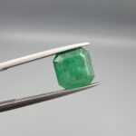 A premium 3.00-carat natural Swat Valley Emerald gemstone held horizontally in stainless steel gemstone tweezers against a smooth, neutral gray background. The loose, earth-mined beryl gem is precision cut into an elegant cushion shape, exhibiting a vivid, saturated medium-to-deep grass-green color with visible natural internal growth features and pristine facet symmetry.