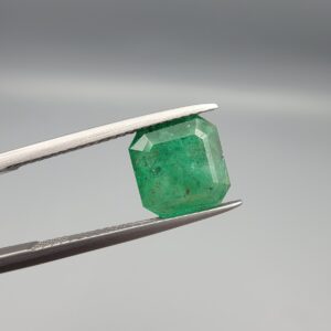 A premium 3.00-carat natural Swat Valley Emerald gemstone held horizontally in stainless steel gemstone tweezers against a smooth, neutral gray background. The loose, earth-mined beryl gem is precision cut into an elegant cushion shape, exhibiting a vivid, saturated medium-to-deep grass-green color with visible natural internal growth features and pristine facet symmetry.