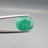 An investment-grade 6.29-carat natural Zambian Emerald gemstone held horizontally in professional metal gemstone tweezers against a smooth, neutral gray studio background. The substantial loose beryl gem is precision cut into an elegant, well-proportioned oval shape, exhibiting a vivid, richly saturated bluish-green color with exceptional transparency and fine facet symmetry.
