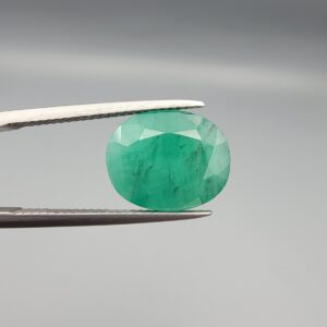 An investment-grade 6.29-carat natural Zambian Emerald gemstone held horizontally in professional metal gemstone tweezers against a smooth, neutral gray studio background. The substantial loose beryl gem is precision cut into an elegant, well-proportioned oval shape, exhibiting a vivid, richly saturated bluish-green color with exceptional transparency and fine facet symmetry.