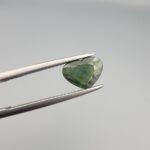 A rare 1.97-carat natural Green Sapphire gemstone held horizontally in professional steel gemstone tweezers against a smooth, neutral gray studio background. The loose Madagascar corundum crystal is masterfully faceted into a romantic heart shape, exhibiting a deeply saturated, sophisticated forest-green body color with fine facet symmetry and crisp meeting lines.