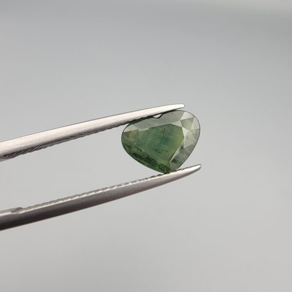 A rare 1.97-carat natural Green Sapphire gemstone held horizontally in professional steel gemstone tweezers against a smooth, neutral gray studio background. The loose Madagascar corundum crystal is masterfully faceted into a romantic heart shape, exhibiting a deeply saturated, sophisticated forest-green body color with fine facet symmetry and crisp meeting lines.