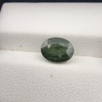 ​A premium 2.96-carat natural Green Sapphire gemstone resting elegantly face-up on a textured white leather jewelry display cushion. The loose Madagascar corundum crystal is precision cut into a symmetrical oval shape, showcasing a deeply saturated forest-green body color with fine facet alignment and sharp crown reflections.