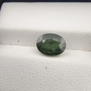 A premium 2.96-carat natural Green Sapphire gemstone resting elegantly face-up on a textured white leather jewelry display cushion. The loose Madagascar corundum crystal is precision cut into a symmetrical oval shape, showcasing a deeply saturated forest-green body color with fine facet alignment and sharp crown reflections.