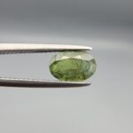 ​A 1.30-carat natural Green Sapphire gemstone held horizontally in professional metal gemstone tweezers against a smooth gray background. The loose Madagascar corundum crystal features a classic faceted oval shape, showcasing an organic olive-green color with dense, microscopic natural internal mineral specks and fine facet symmetry.