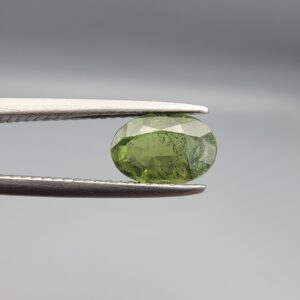 A 1.30-carat natural Green Sapphire gemstone held horizontally in professional metal gemstone tweezers against a smooth gray background. The loose Madagascar corundum crystal features a classic faceted oval shape, showcasing an organic olive-green color with dense, microscopic natural internal mineral specks and fine facet symmetry.