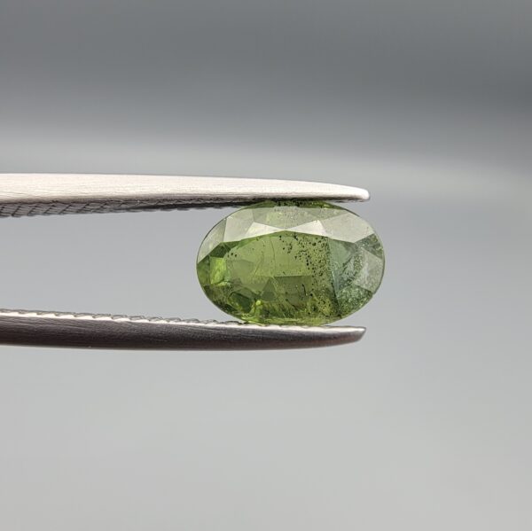 ​A 1.30-carat natural Green Sapphire gemstone held horizontally in professional metal gemstone tweezers against a smooth gray background. The loose Madagascar corundum crystal features a classic faceted oval shape, showcasing an organic olive-green color with dense, microscopic natural internal mineral specks and fine facet symmetry.