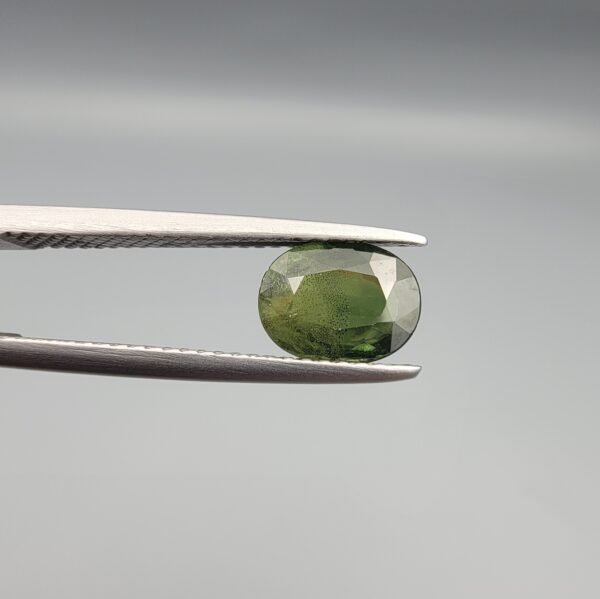 ​A premium, beautifully faceted 1.63-carat natural Green Sapphire gemstone held securely in metal tweezers against a clean, neutral grey background. The loose corundum gem showcases an expert oval cut with a well-proportioned table, displaying a rich, uniform olive-green color with fine, genuine internal crystal characteristics that verify its earth-mined origin.