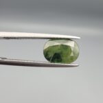 ​A premium, beautifully faceted 3.17-carat natural Green Sapphire gemstone held securely in metal tweezers against a clean, neutral grey background. The loose corundum gem showcases an expert oval cut with a well-proportioned table, displaying a rich, uniform olive-green color with fine, genuine internal crystal characteristics that verify its earth-mined origin.