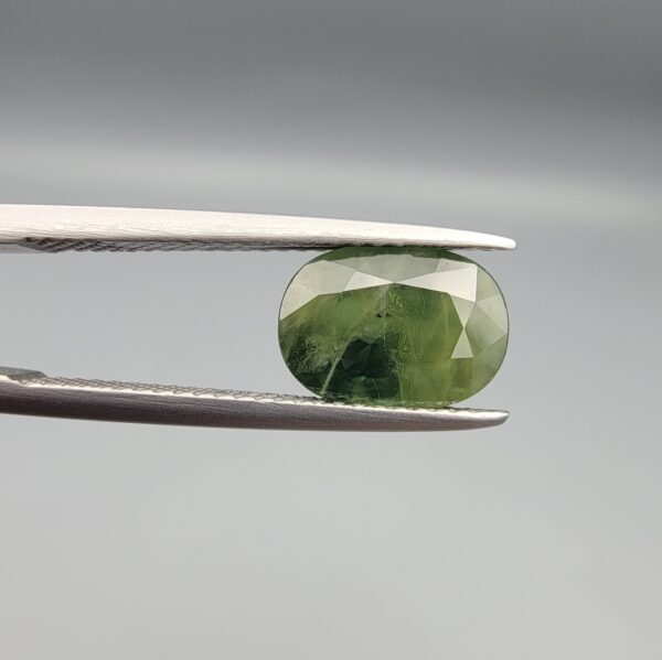 ​A premium, beautifully faceted 3.17-carat natural Green Sapphire gemstone held securely in metal tweezers against a clean, neutral grey background. The loose corundum gem showcases an expert oval cut with a well-proportioned table, displaying a rich, uniform olive-green color with fine, genuine internal crystal characteristics that verify its earth-mined origin.